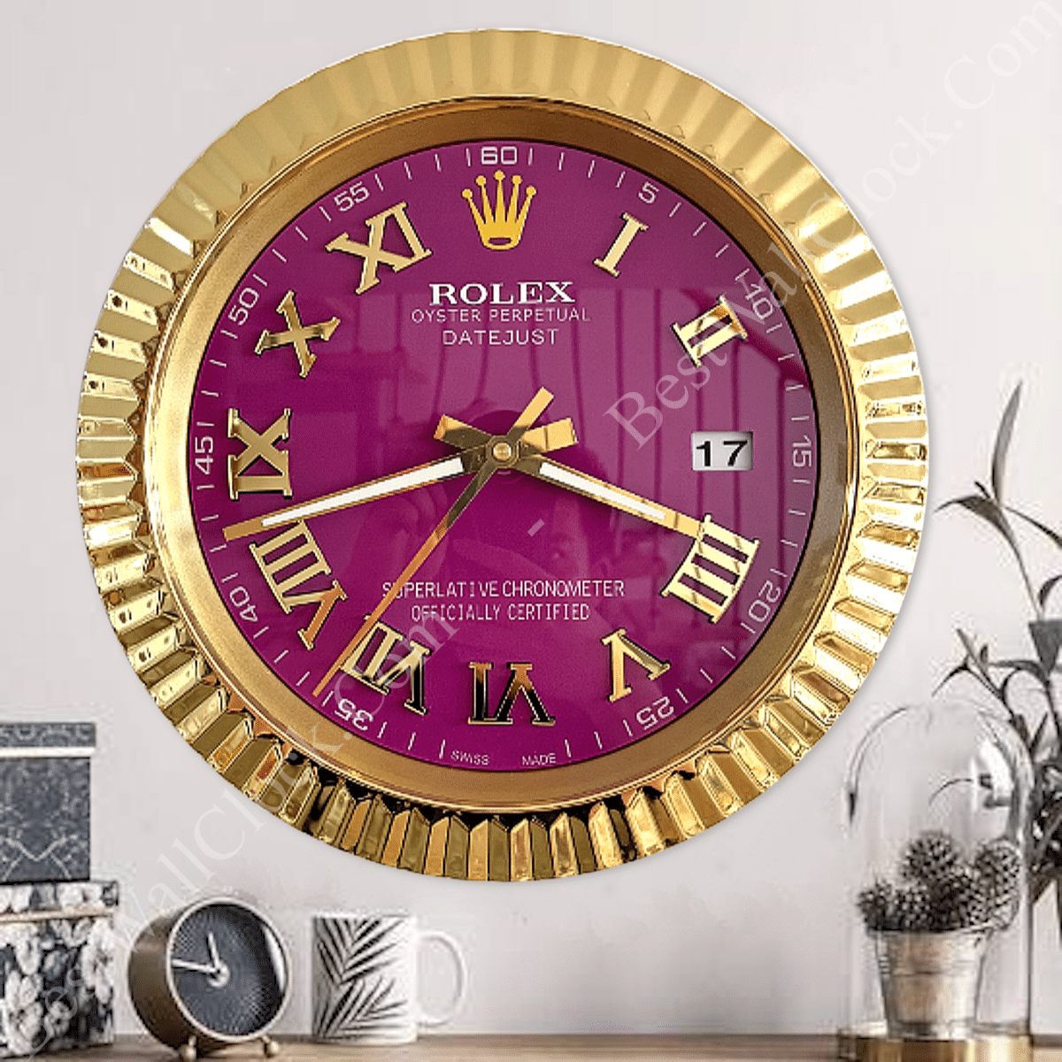 Gold-framed wall clock with a purple face featuring large gold Roman numerals and the brand name Rolex, displaying the time as 8:15 with a date window showing the 17th, mounted on a light grey wall above a wooden surface with a small black clock, a white mug with leaf design, and a glass cloche with pine cones and a green plant.