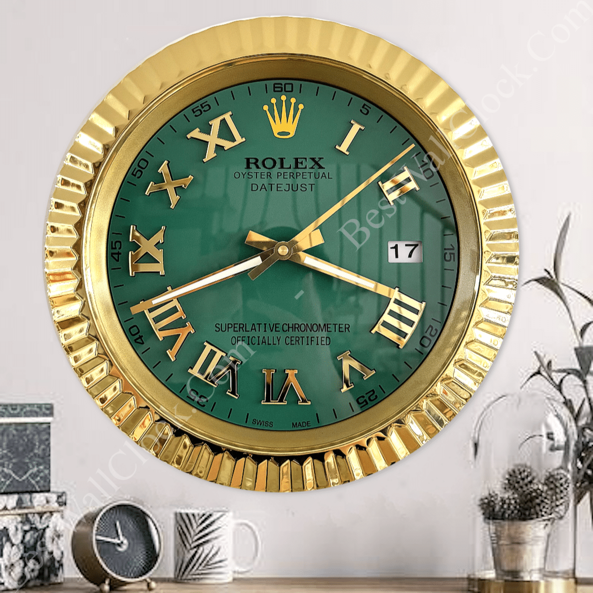 Gold-framed Rolex Oyster Perpetual Datejust wall clock with a green face, gold Roman numerals, and date display set to 17, hanging on a white wall over a table with a small black clock, a white mug with a leaf design, and a plant under a glass dome.