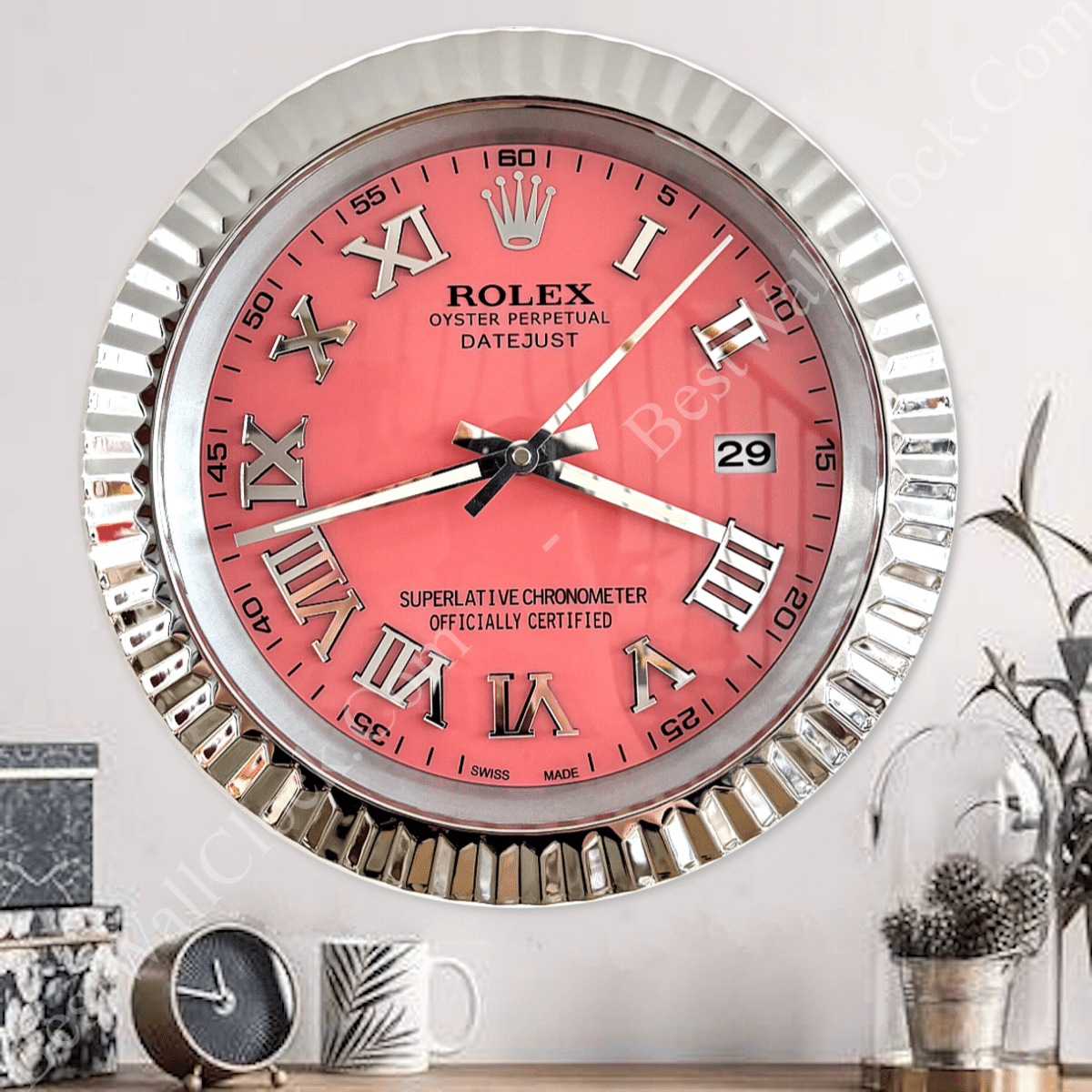 Wall clock with a shiny silver frame, coral pink face, large silver Roman numerals, and black hour and minute hands, displaying time as 8:17 and date as 29, with decorative items on a wooden surface below and a white wall background