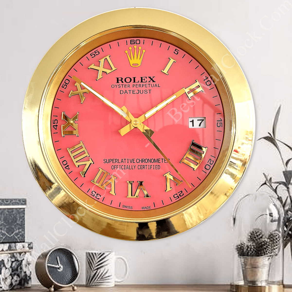 Golden-framed wall clock with a red face and Roman numerals, showing the time as 10:53 and date 17, mounted above a wooden surface with a small black clock, a white mug, and two glass containers with plants and pinecones on a light-coloured wall.
