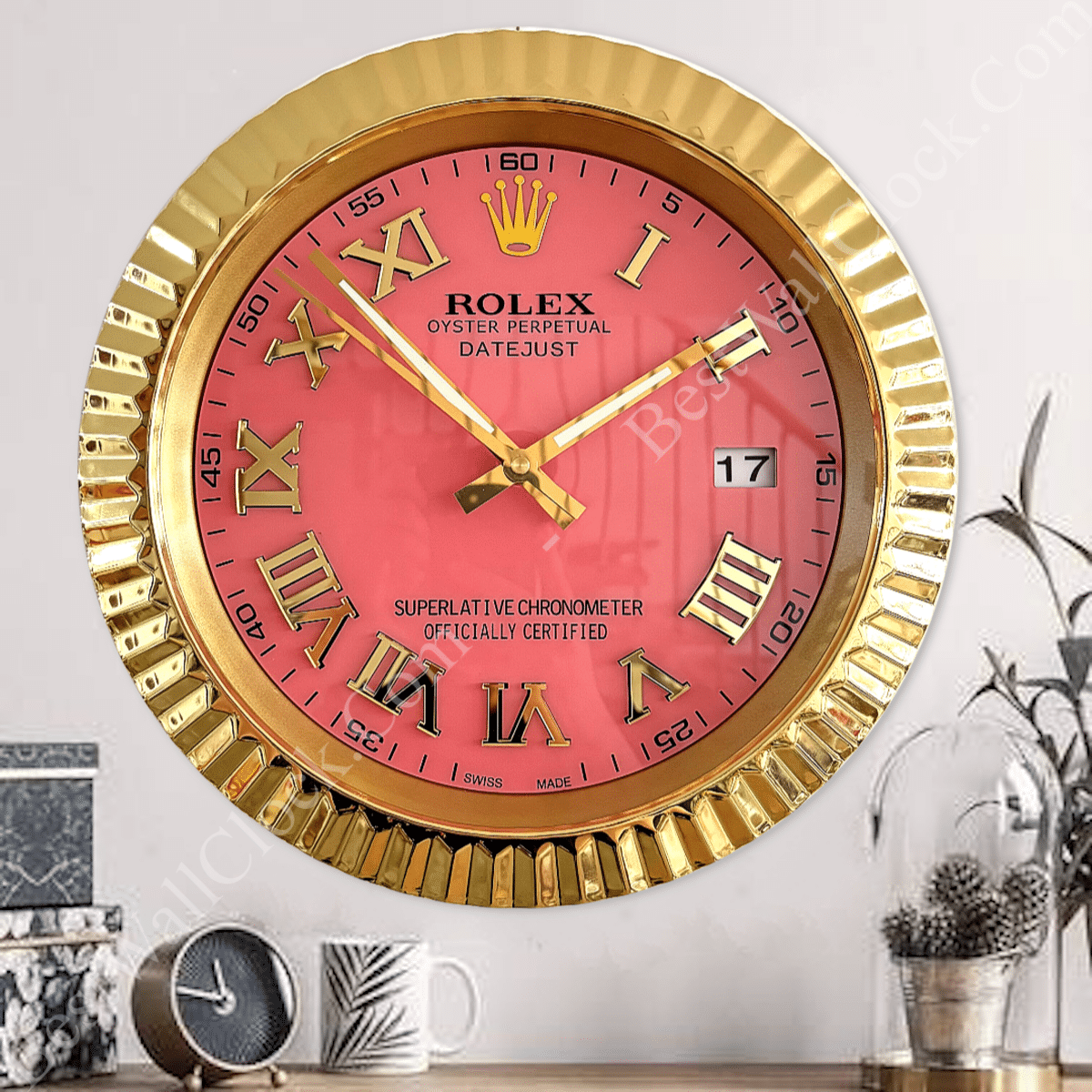 Round wall clock with a gold ridged frame, pink face, and gold Roman numeral hour markers, showing the time as approximately 10:56, with the date displayed as 17; placed above a shelf with a small clock, a plant-patterned mug, and decorative plants under a glass dome.