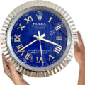 Blue Rolex Oyster Perpetual Datejust wall clock with silver Roman numerals and hands, held by two hands against a white background