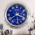 Wall clock designed like a Rolex Datejust watch with a royal blue face, silver Roman numerals, date window at three o'clock, encased in a fluted silver bezel on a white wall above a shelf holding a smaller clock, a mug, and a small plant in a glass container.