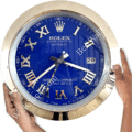 Close-up of a round Rolex wall clock with a blue face, silver Roman numerals, and a silver frame, showing the time as approximately 7:16, held by two hands on either side.