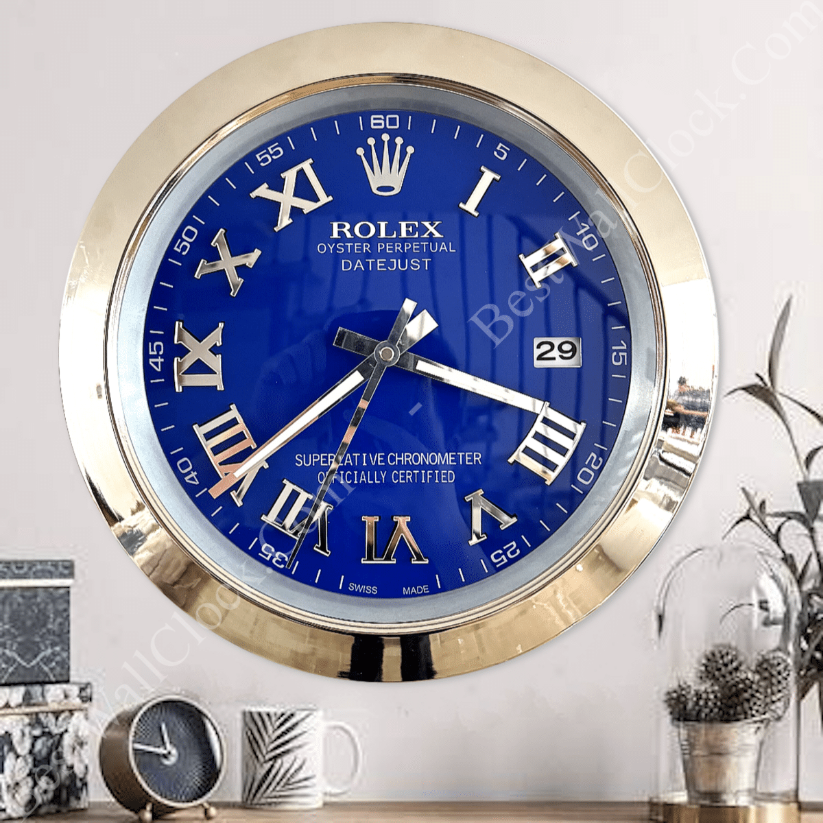 Round wall clock styled like a Rolex Datejust watch with a blue face, gold Roman numerals, date window at the 3 o'clock position, and a gold rim, mounted on a white wall above a surface with a small clock, a mug, and decorative items including plants and a glass dome with pine cones.