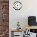 Modern office corner with a black leather chair, wooden desk, side table with potted plants and books, and a round wall clock with a silver frame and Roman numerals on a white wall next to exposed brick.