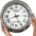 Close-up of a round wall clock designed like a Rolex Oyster Perpetual Datejust watch face with Roman numerals and a date window showing the 28th, held by two hands on either side against a white background.