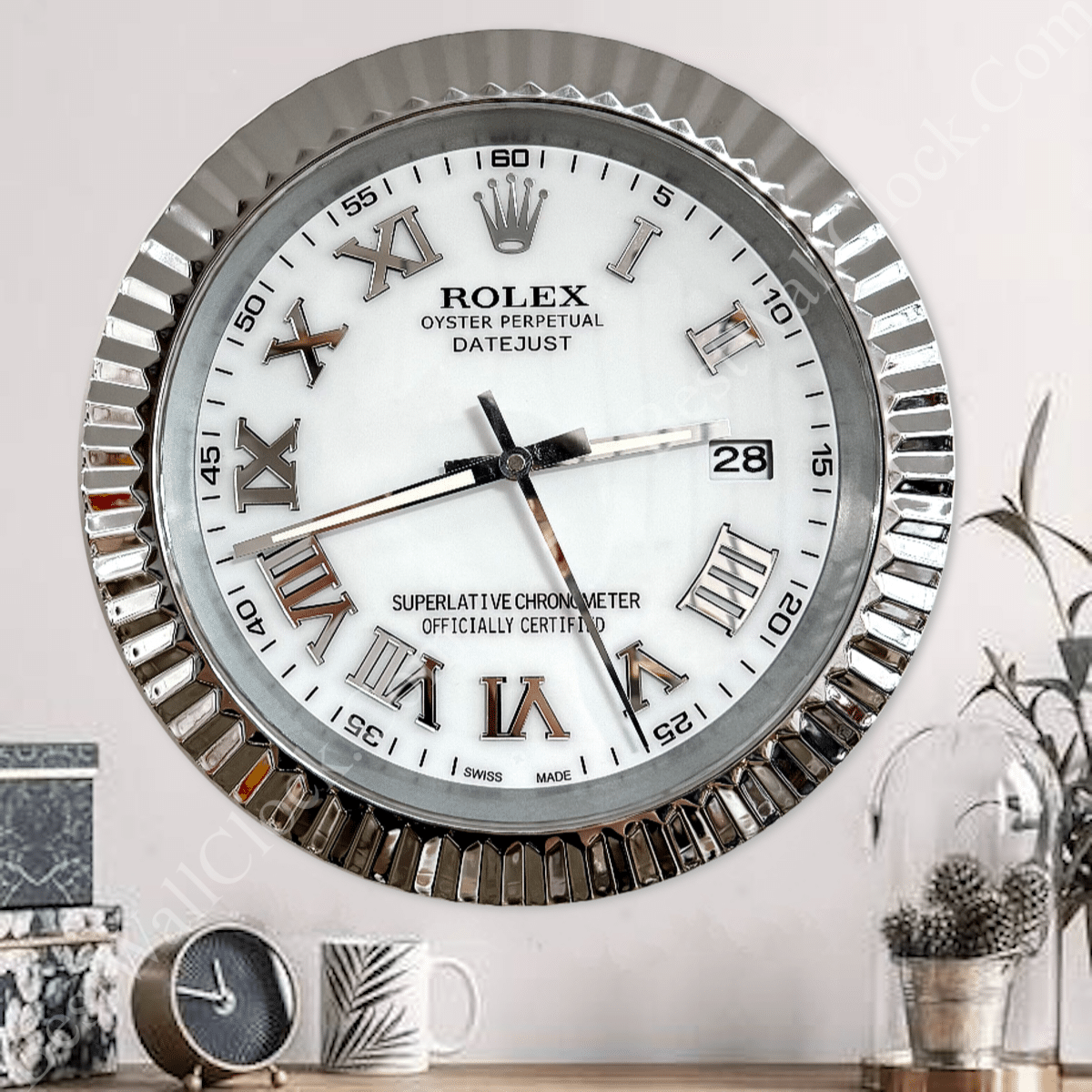 Close-up of a silver Rolex wall clock with roman numerals and a fluted bezel, mounted on a light wall above a wooden surface holding a small round clock, a white mug with a leaf pattern, and a glass-covered plant pot with pine cones and greenery.