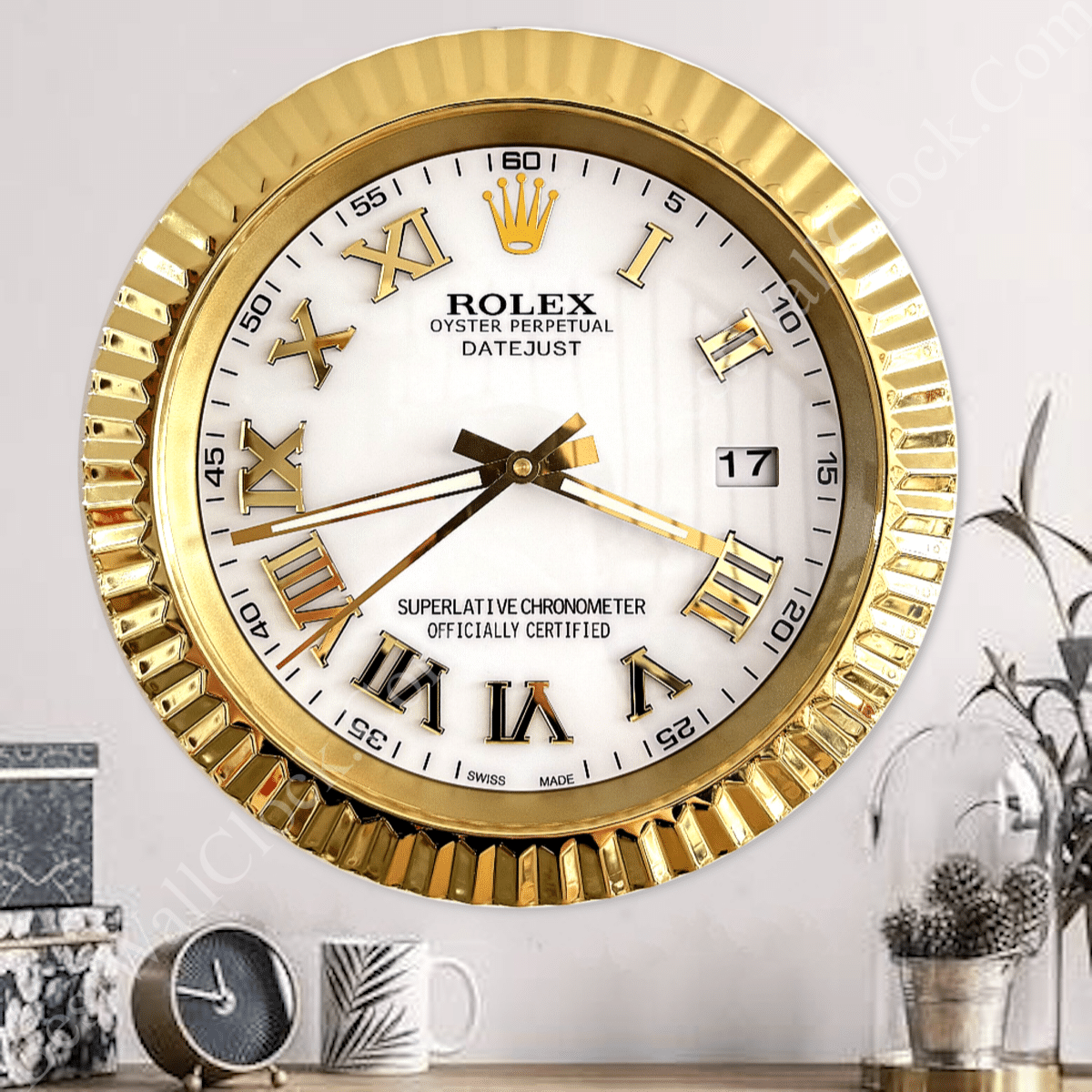 Gold-framed Rolex Oyster Perpetual Datejust wall clock with white face, gold Roman numerals, date window at 3 o'clock, and gold hour, minute, and second hands, mounted on a light wall above a table with a small round clock, coffee mug, and decorative plant under a glass dome.