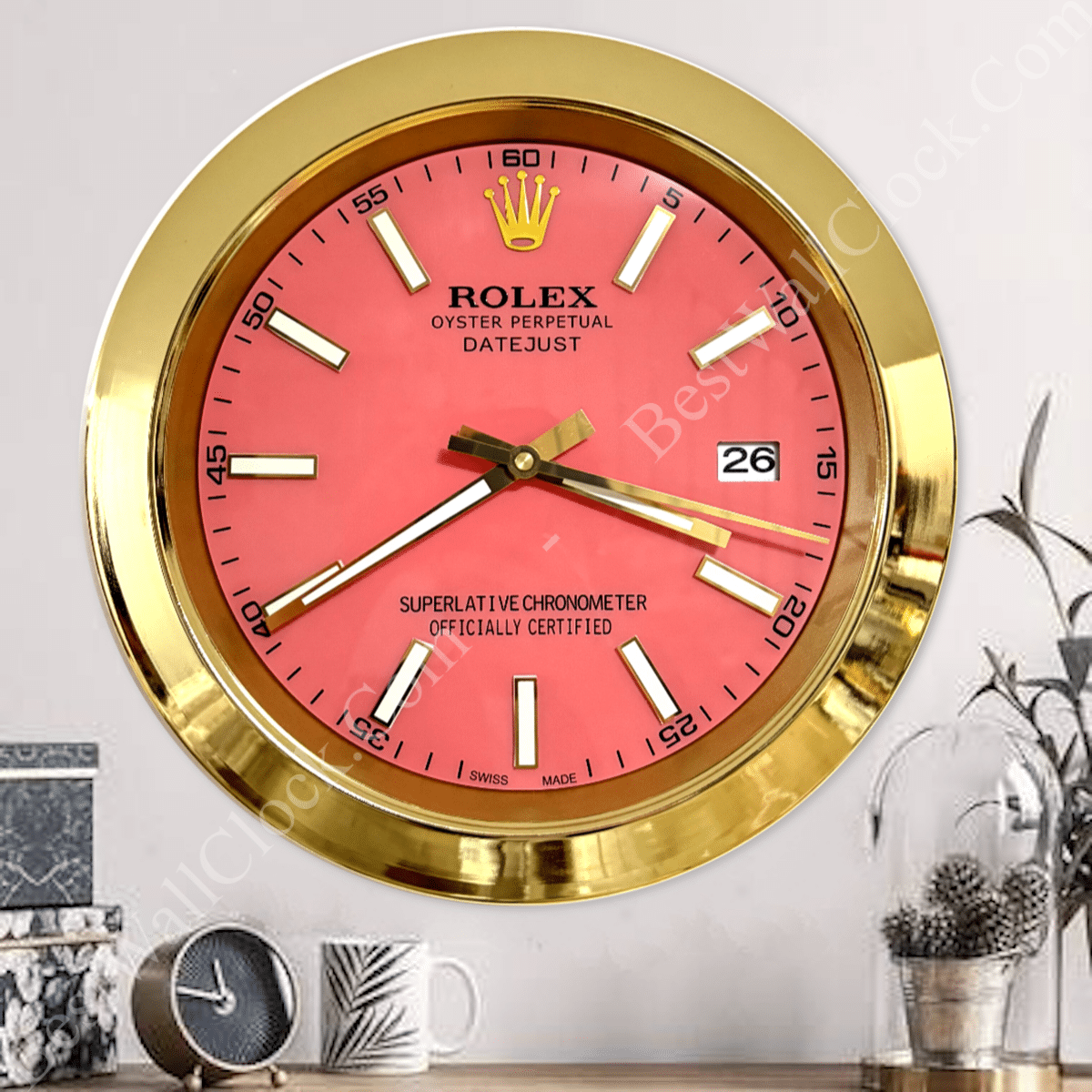 Wall clock designed to resemble a Rolex watch with a gold frame, pink face, white and gold hour markers, and hands showing the time as 8:19 and the date as 26, placed on a white wall above a table with an alarm clock, mug, and decorative plants.