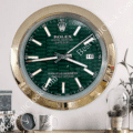 Wall clock designed to resemble a Rolex Datejust watch face with a green patterned dial, gold-tone bezel, and white hour markers, showing the time as 3:45 and the date as 16, mounted on a wall with decorative items below including a clock, a mug, and a plant in a glass dome.