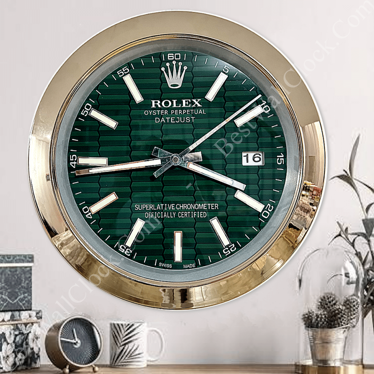 Wall clock designed to resemble a Rolex Datejust watch face with a green patterned dial, gold-tone bezel, and white hour markers, showing the time as 3:45 and the date as 16, mounted on a wall with decorative items below including a clock, a mug, and a plant in a glass dome.
