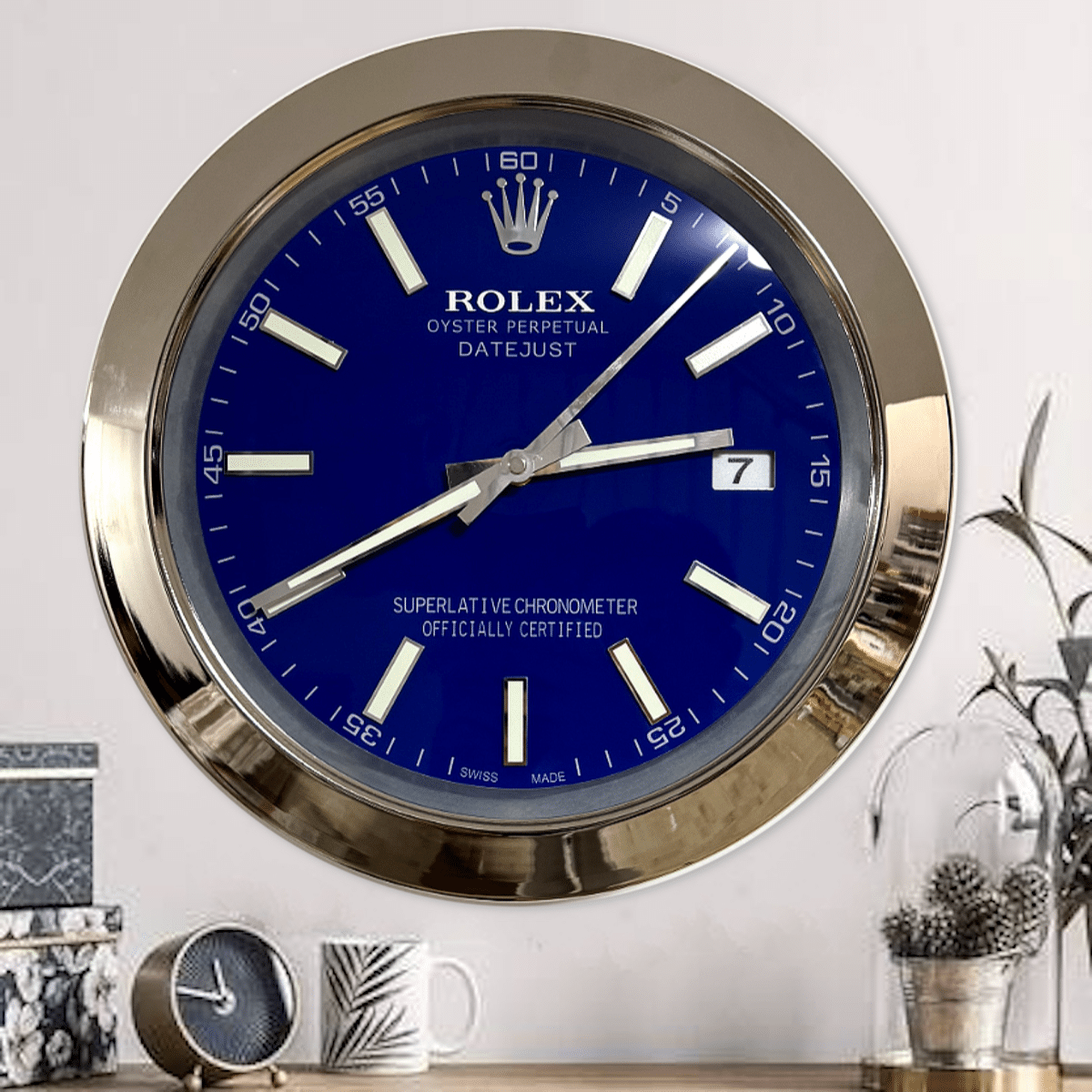 Wall clock designed to resemble a Rolex Oyster Perpetual Datejust watch with a blue face, white hour markers, silver hands, and a gold bezel, mounted above a desk with a small round clock, a white mug with a leaf design, and decorative plants in glass containers.