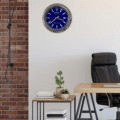 Modern office corner with a black leather chair, wooden desk, small stacked tables holding books and potted plants, a brick wall on the left, and a blue-faced Rolex wall clock showing 3:39 on a plain light wall.