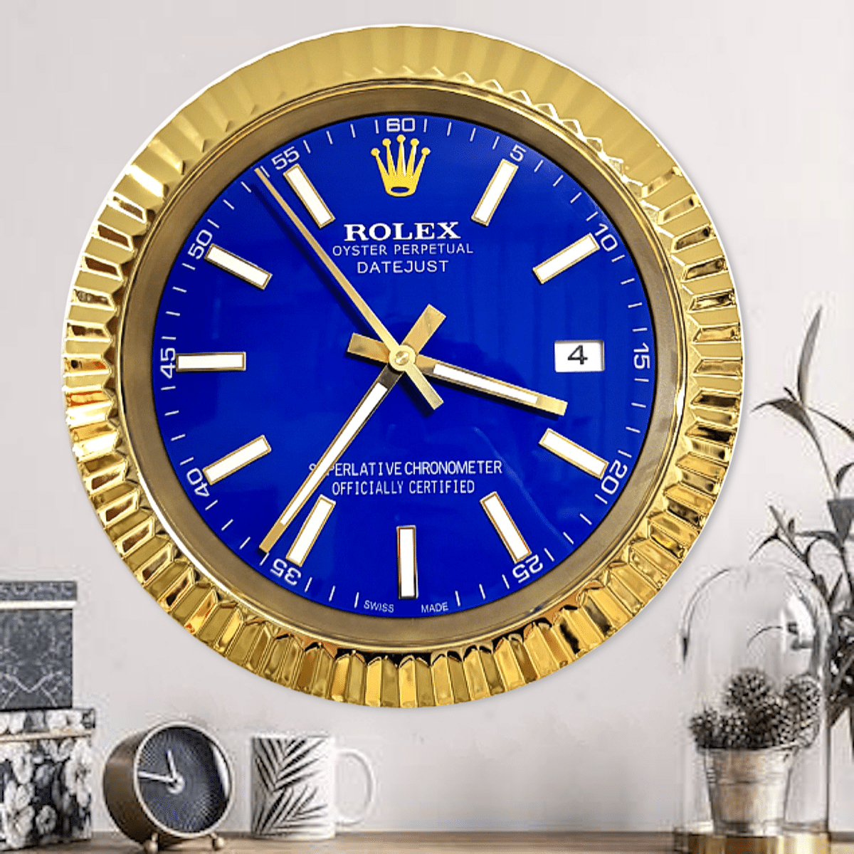 Gold-framed Rolex wall clock with a blue face, white hour markers, and gold hands showing the time as 3:38, placed above a table with a small round clock, a white mug, and a glass plant terrarium on a light grey wall background.