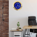 Modern office corner with a wooden desk, black leather chair, side table with potted plants and stacked books, and a gold-framed blue Rolex wall clock showing the time as 4:35:23 on a white wall next to a red brick wall.