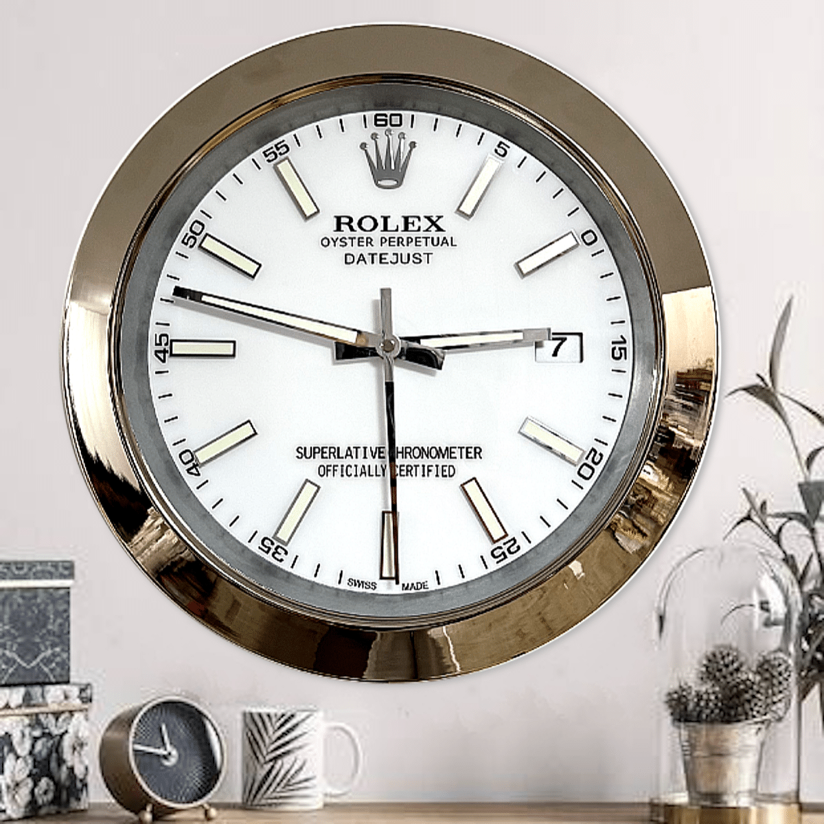 Round wall clock designed like a Rolex watch with a gold rim, white face, black markings, and a date window showing the number 7, hanging above a table with a small clock, a mug with a leaf pattern, and a glass dome covering pine cones.