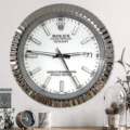 Wall-mounted Rolex Datejust clock with a silver fluted bezel and white face showing the time as 03:46:26, above a wooden surface with a small clock, a patterned mug, and decorative plants under a glass dome.