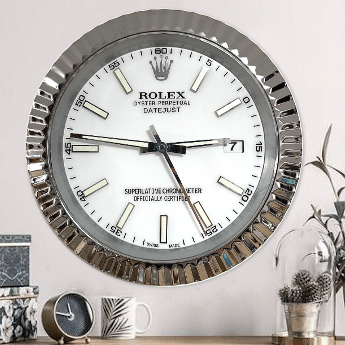 Wall-mounted Rolex Datejust clock with a silver fluted bezel and white face showing the time as 03:46:26, above a wooden surface with a small clock, a patterned mug, and decorative plants under a glass dome.