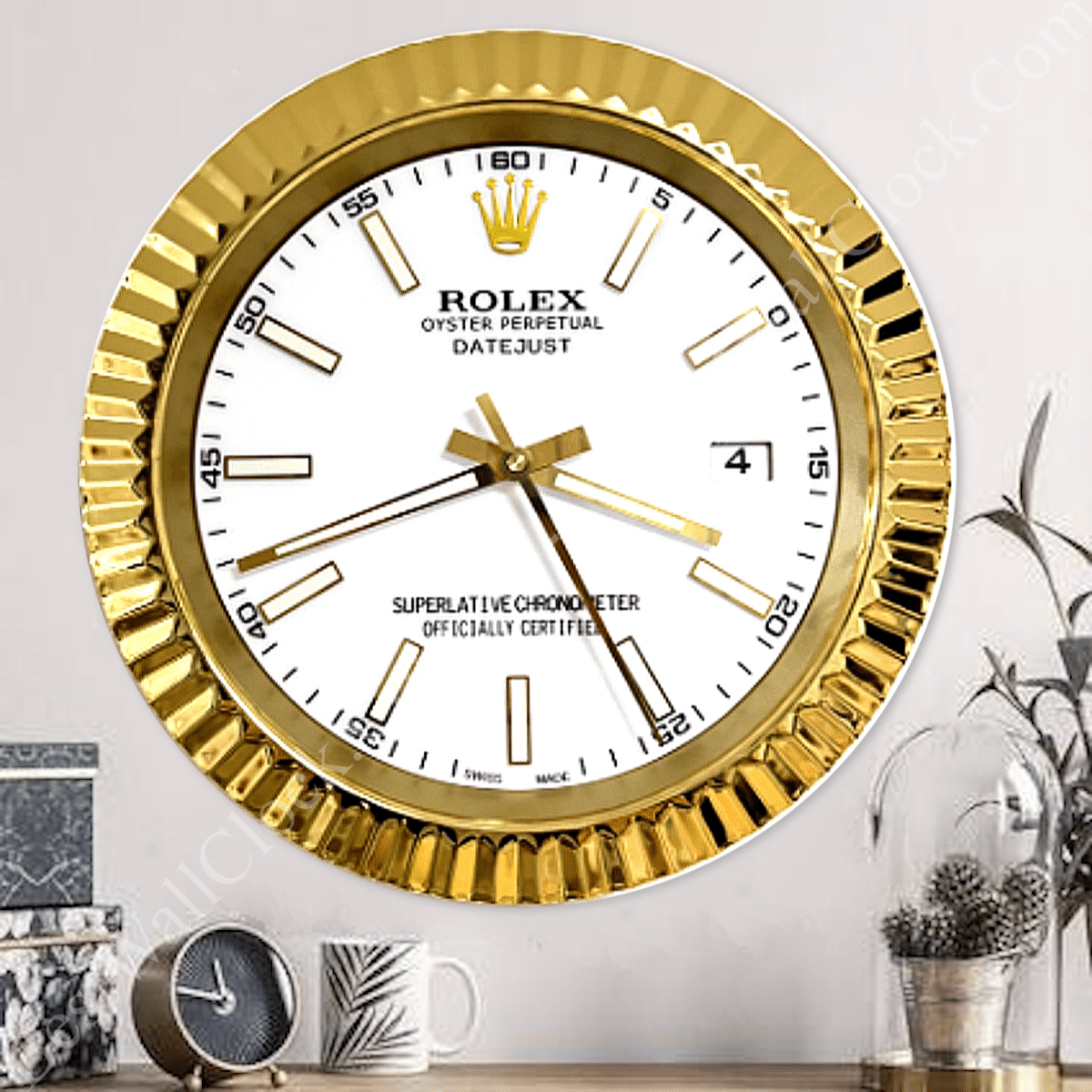 Wall clock designed like a gold Rolex watch face with white background and gold hour markers, mounted on a white wall above a wooden surface with a small round clock, a white mug with a leaf pattern, and a glass dome with pine cones inside beside a plant.