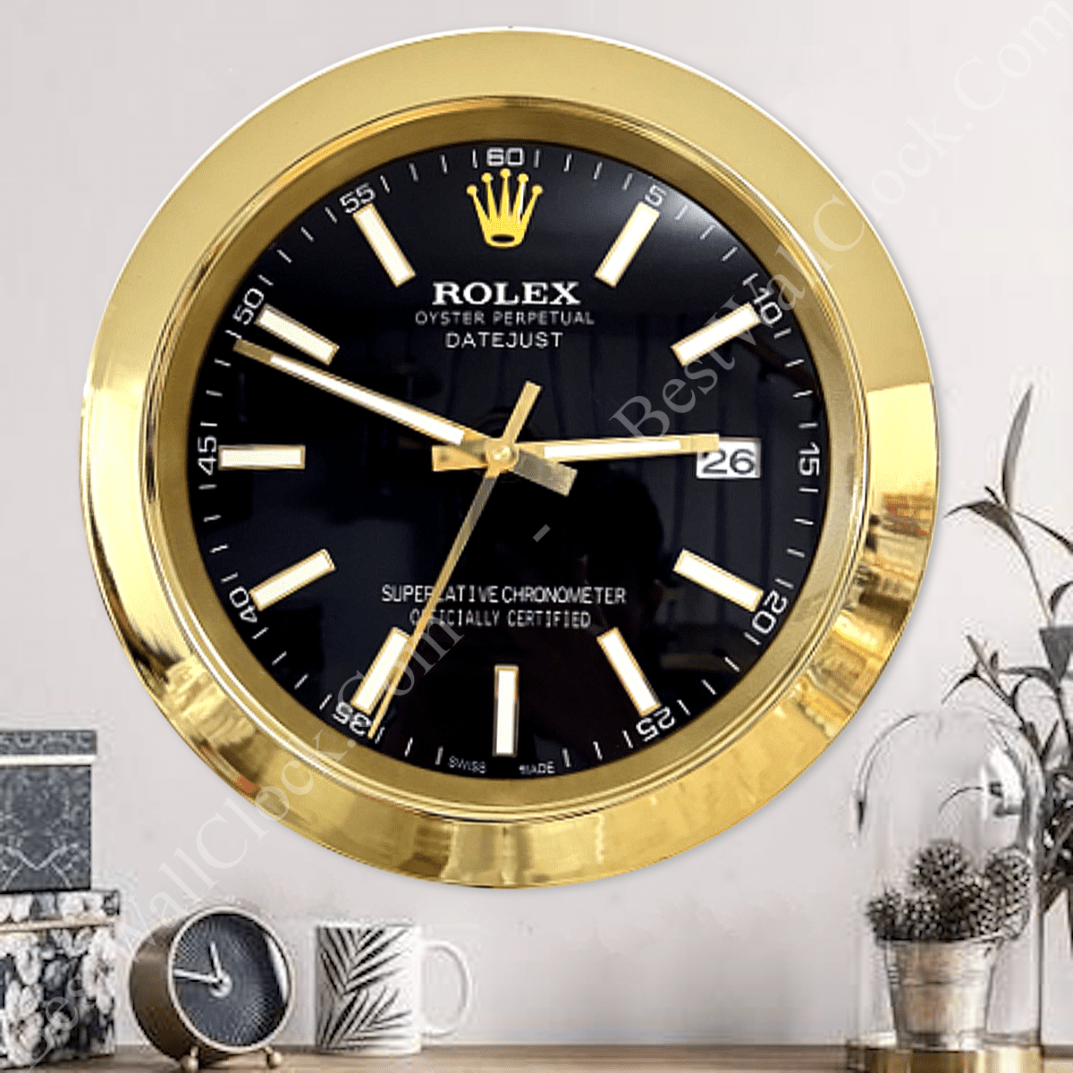 Wall clock styled like a gold Rolex Datejust watch with a black face, gold hour markers, and a date display showing 26, mounted on a light wall above a table with a small round clock, a white mug with a leaf design, and a plant in a glass cloche.