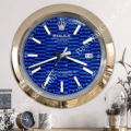 Wall clock designed like a Rolex Oyster Perpetual Datejust watch with a blue patterned face, gold bezel, white hour markers, and date display showing 16, mounted on a white wall above a shelf with a clock, cup, and plant.