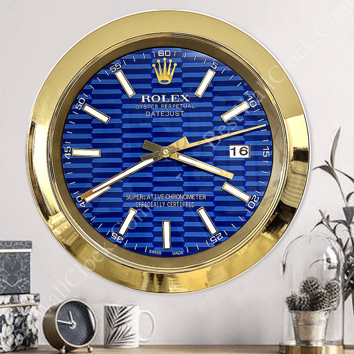 Wall clock styled like a Rolex watch face with a blue textured background, gold rim, and gold hour, minute, and second hands, showing the date as 16, mounted on a white wall above a shelf with decorative items including a small clock, a cup, and a plant under a glass dome.