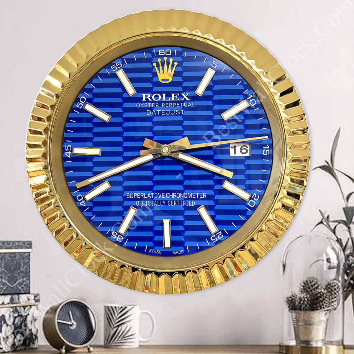 Wall clock designed to resemble a Rolex wristwatch, featuring a gold-tone fluted bezel, deep blue textured dial with gold and white hour markers, three hands, and a date display showing the 16th, mounted on a light wall above a wooden surface with decorative items.
