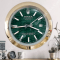 Wall clock designed to resemble a Rolex Oyster Perpetual Datejust watch with a green honeycomb-patterned face, gold outer rim, white hour markers, and a date display showing the number 16, mounted on a light-colored wall above a shelf with decorative items including a small clock, mug, and plant.