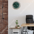 Modern office space with a black leather office chair, wooden desk, side table holding stacked books and two small potted plants, and a wall clock resembling a Rolex watch with a green face and silver bezel mounted on a white wall next to an exposed brick wall with a hanging cable.