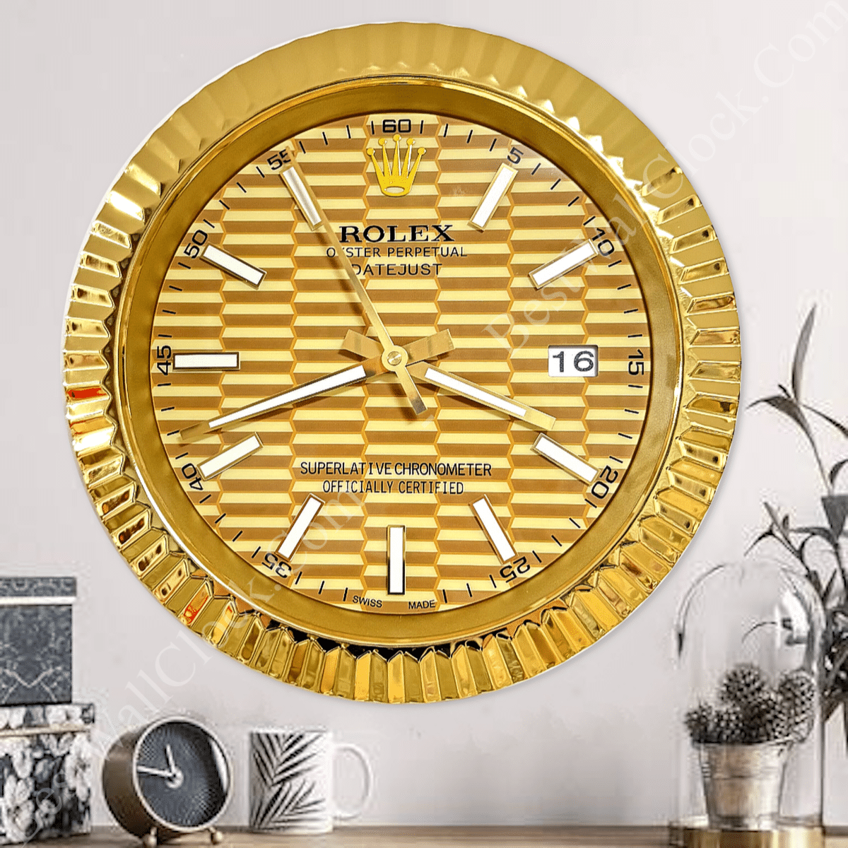 A large wall clock designed like a Rolex Datejust watch face with a gold fluted bezel and a textured gold dial, displaying the time as 3:41:56 and a date window showing 16, mounted on a white wall above a wooden surface with a small black and gold clock, a white mug with a leaf design, and a potted plant under a glass dome.