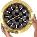 Rolex well clock showing black and grey striped face with gold bezel, gold hour markers, and date display at 3 o'clock, held by two hands.