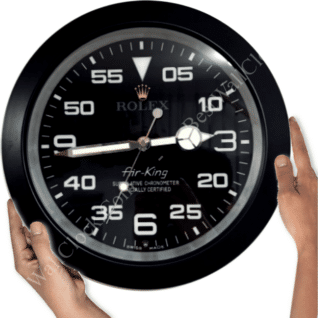 A black Rolex Air-King wall clock with white numerals and hands, held by two hands on either side, showing the time as approximately 3:44.