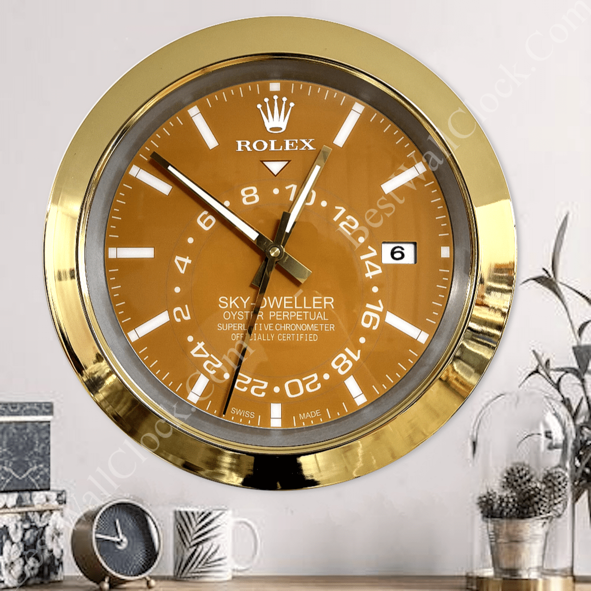 Gold-framed Rolex Sky-Dweller wall clock with brown face, white hour markers, and date display reading 6, mounted on a white wall above a wooden surface with a small round clock, a white mug with leaf pattern, and a plant in a glass pot.