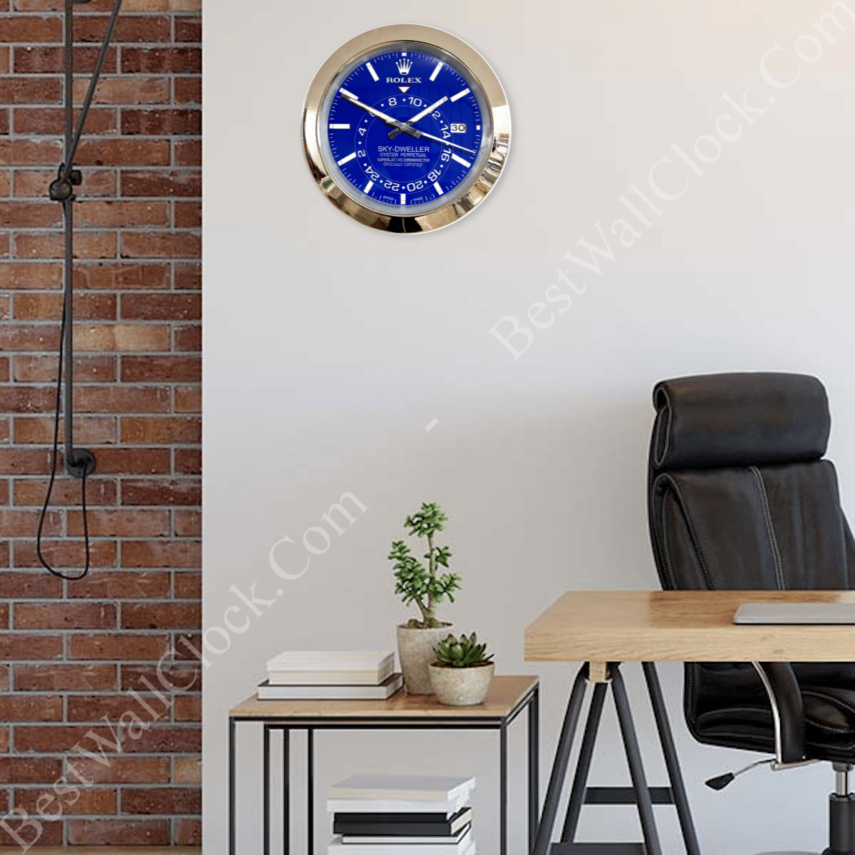Modern office interior with a black leather office chair, a wooden desk, a small table with stacked books and two small potted plants, and a blue Rolex Sky-Dweller wall clock mounted on a white wall next to a brick wall with a black wall-mounted lamp.