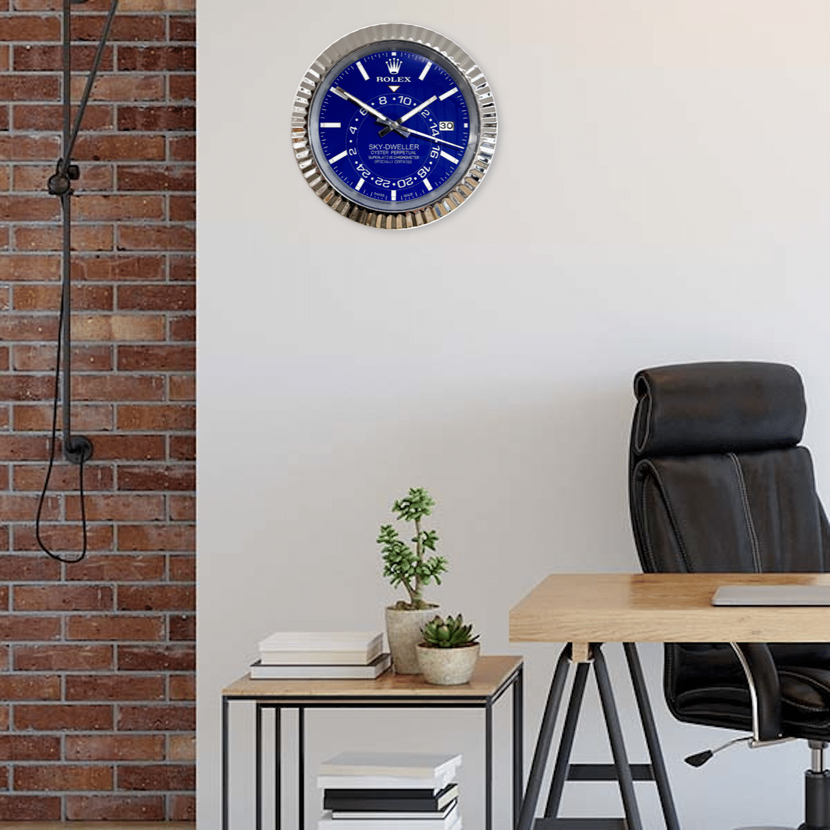 Modern office corner with a black leather chair, light wooden desk, small side table featuring two potted plants and stacked books, and a wall clock designed to resemble a Rolex watch with a blue face and silver frame mounted on a white wall next to a brick wall.