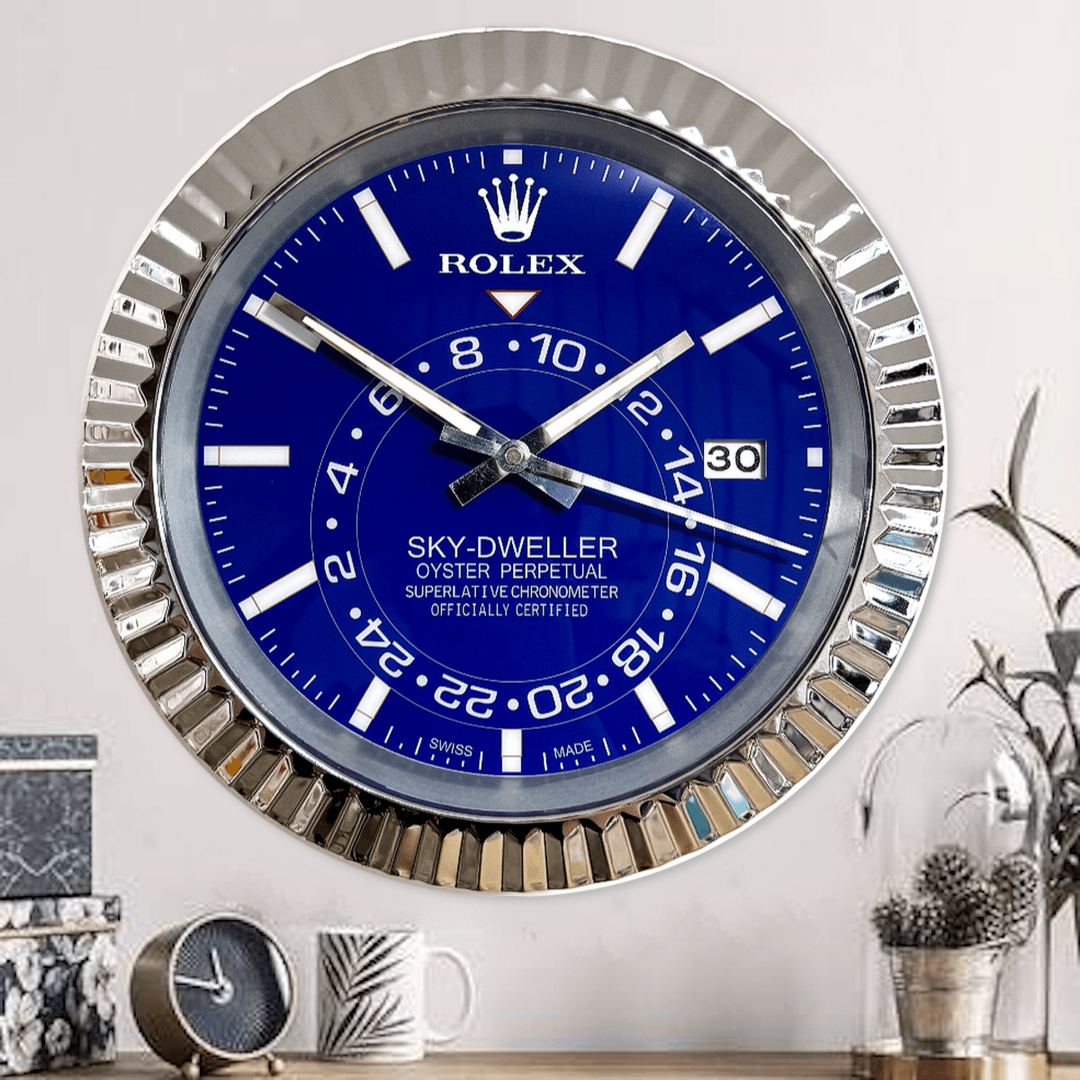 Wall clock designed to resemble a Rolex Sky-Dweller watch with a metallic fluted bezel and a blue dial showing the time 9:53 with a date display of 30, mounted on a wall above a wooden surface with a small clock, a mug, and a decorative plant.