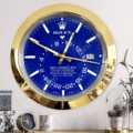 Wall clock designed like a Rolex Sky-Dweller watch with a gold frame and a blue face, showing the date 30, hanging above a table with a small clock, a white mug with a leaf pattern, and a glass dome containing pine cones and a plant.