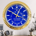 Wall clock designed like a Rolex Sky-Dweller watch with a blue face, gold frame, and white hour markers hanging on a wall above a wooden surface with a small clock, a leaf-patterned mug, and a plant in a glass dome.