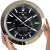 Close-up of a large Rolex Sky-Dweller wall clock with a black dial, white hour markers, and silver hands, held by two hands, showing the time as approximately 10:08:23.