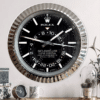 A large wall clock designed to resemble a Rolex Sky-Dweller watch with a black dial, silver hour markers, and a fluted silver bezel, mounted on a light-coloured wall above a wooden surface with decorative items including a small clock, a mug, and a plant under glass domes.