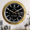 Wall clock designed to look like a Rolex Sky-Dweller watch face with a black dial, white hour markers, gold hands, and a gold ridged bezel, mounted on a light-colored wall above a wooden surface with decorative items including a small clock, mug, and glass plant terrarium.
