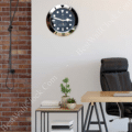 Modern office space with a wooden desk, black leather office chair, small wooden side table with potted plants and books, exposed brick wall on the left, and a round wall clock resembling a Rolex Yacht-Master watch on a plain light-coloured wall