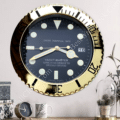 Large wall clock designed to resemble a gold luxury watch face with black dial, luminous hour markers and hands, displaying the date as 16, mounted on a pale grey wall above a wooden surface with a small round table clock, a white patterned mug, and a glass cloche containing a potted plant.