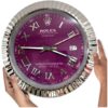 Hands holding a round wall clock with a metallic fluted bezel, a purple dial, silver Roman numerals, and silver hands showing the time as 09:17 with a date window displaying 27; the clock face features Rolex branding and text.