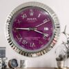 Wall-mounted Rolex Oyster Perpetual Datejust-style clock with a purple face, silver Roman numerals, and silver fluted rim, showing the time as 3:45 and the date as 27, above a desk with a small clock, mug, and decorative objects.