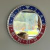 Round wall mirror with a blue and red numbered bezel resembling a GMT watch bezel, reflecting a living room with a brown pillow on a beige chair and a view of a garden with a pool and greenery outside a window.