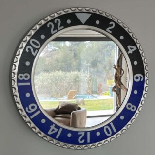 Circular wall mirror resembling a watch bezel with alternating black and blue segments marked with numbers from 2 to 22, reflecting a living room scene with a chair, brown pillows, and a window showing a garden with trees, lawn, a pool, and an outdoor lounge chair.