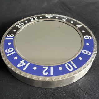 Round wall clock styled like a watch bezel with a silver frame, black and blue numbered segments, and a blank reflective face without hands or numbers.