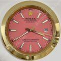 Wall clock styled as a Rolex Oyster Perpetual Datejust watch with a pink face, gold rim, white hour markers, and date display showing 26, with the time showing 7:39:15.